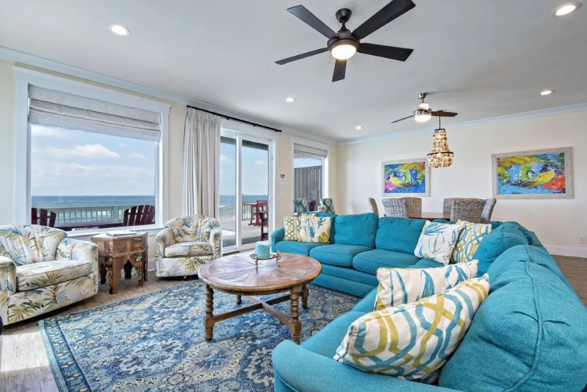 A cheerful, coastal-inspired living area featuring a large turquoise sectional, round wooden table, colorful artwork, and an ocean view through oversized windows. The room is bright and airy, creating an inviting space for both relaxation and group activities.