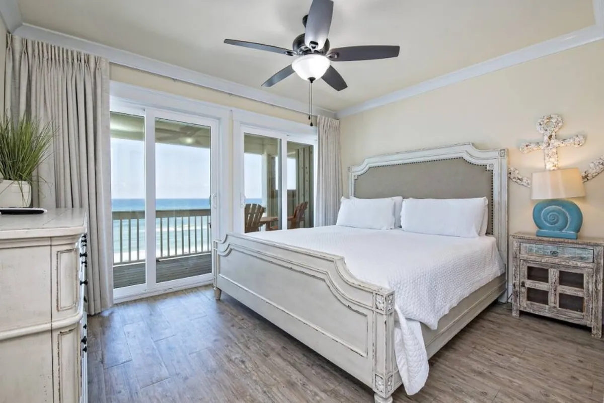 A king-size bed sits in a serene, sunlit room with a view of the balcony and ocean beyond. Decorative lamps and warm wood tones create an inviting atmosphere, perfect for restful nights and peaceful mornings.