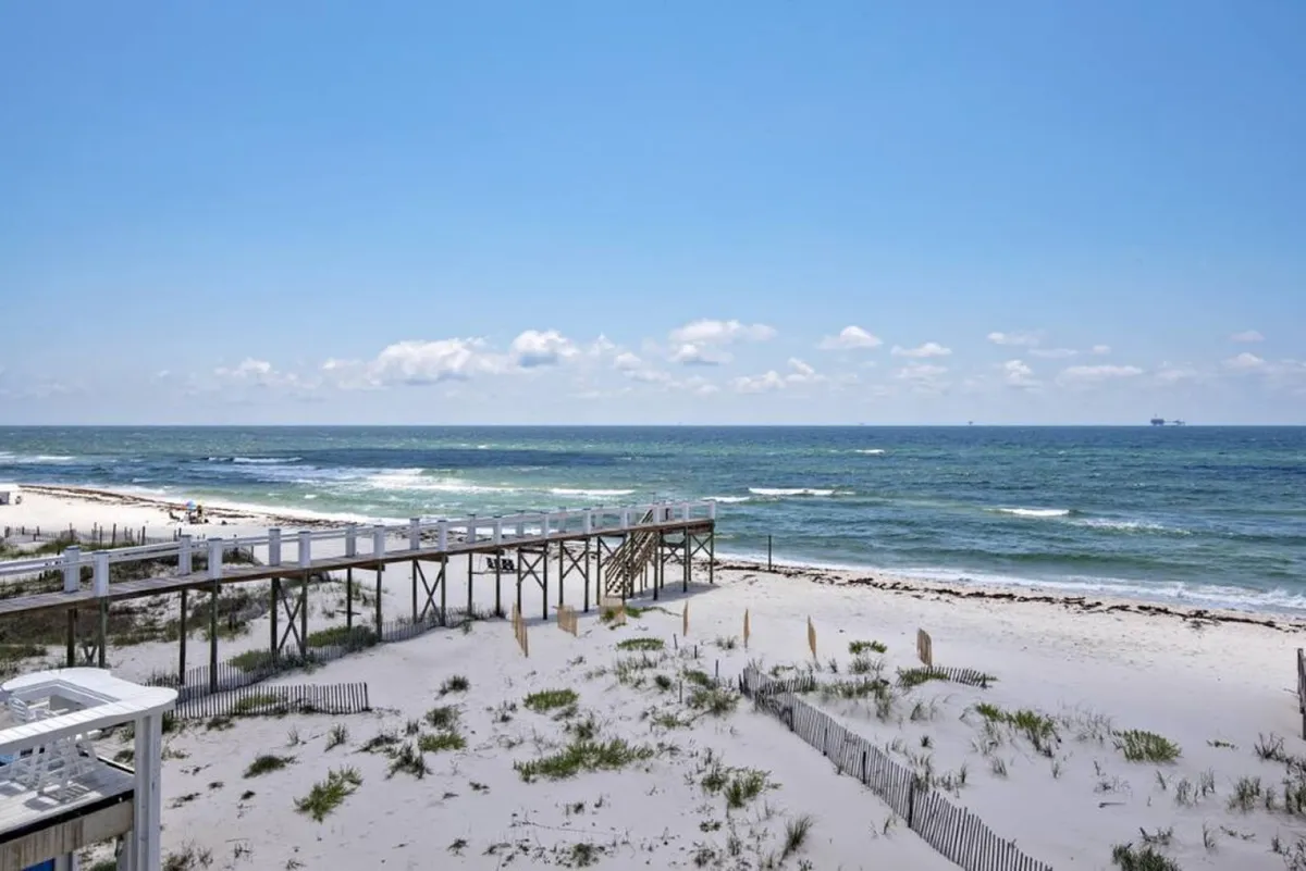A sandy boardwalk winds through dune grass, leading directly to a secluded stretch of white sand beach and expansive ocean beyond. The path feels private and inviting, promising easy access to the water and an escape from the crowds. The natural elements and sense of openness create a tranquil vibe.