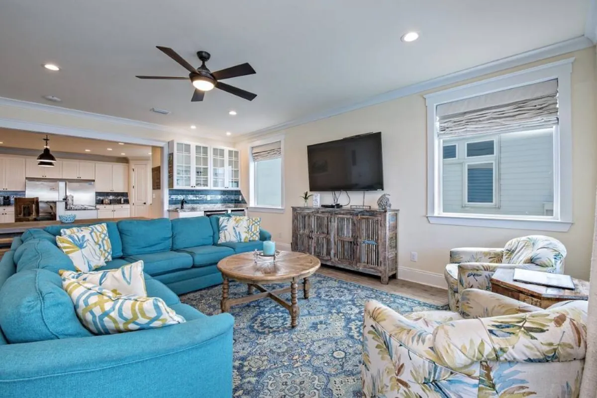 The image features a large teal sectional sofa, a wooden coffee table, floral armchairs, and a TV mounted on the wall, all set within a welcoming living room. Generous windows allow sunlight to stream in, creating a space designed for both relaxation and group interaction.