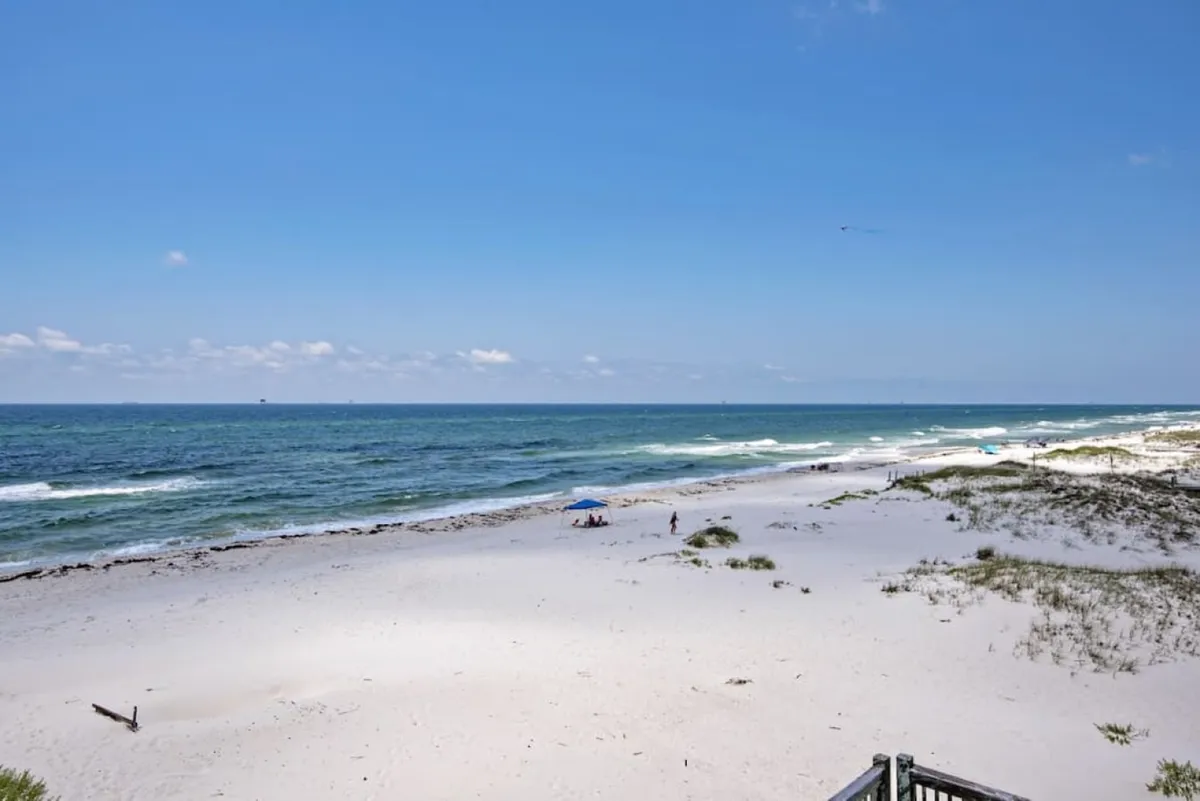 The image shows a tranquil beach scene with the ocean stretching out to the horizon, sand dunes in the foreground, and people enjoying the beach. The expansive sky and relaxed vibe highlight the natural beauty of Gulf Shores.