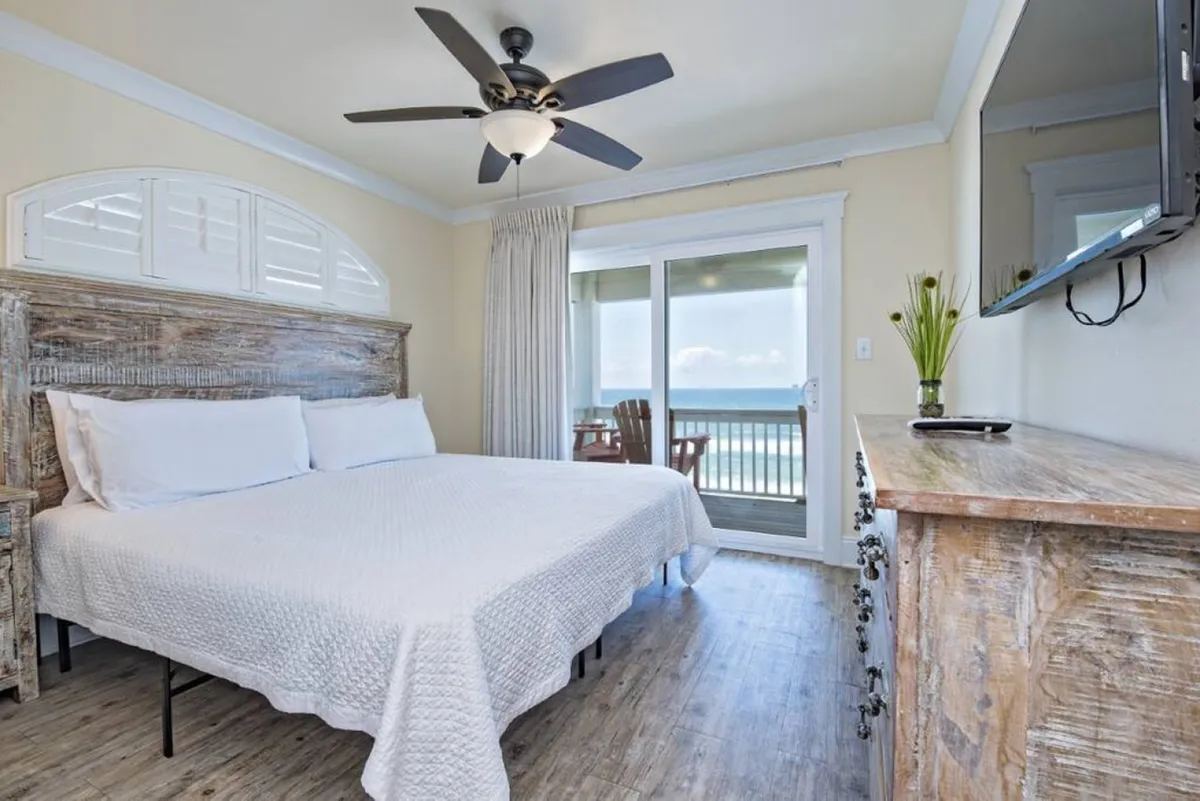 A spacious king-size bed faces a sliding glass door that opens onto a balcony, with wooden furniture and a ceiling fan completing the tranquil bedroom. The room is filled with natural light and offers unobstructed views of the Gulf, inviting guests to relax or enjoy coffee while soaking in the scenery.