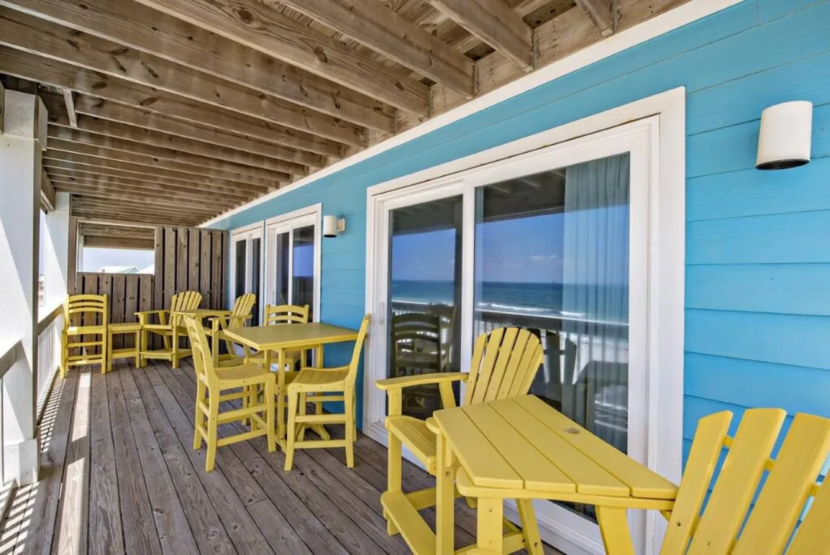 The image features a vibrant exterior seating area with bright yellow chairs and a blue wall, overlooking a panoramic sea view. The setup is casual yet inviting, ideal for group relaxation, breakout sessions, or informal meetings.