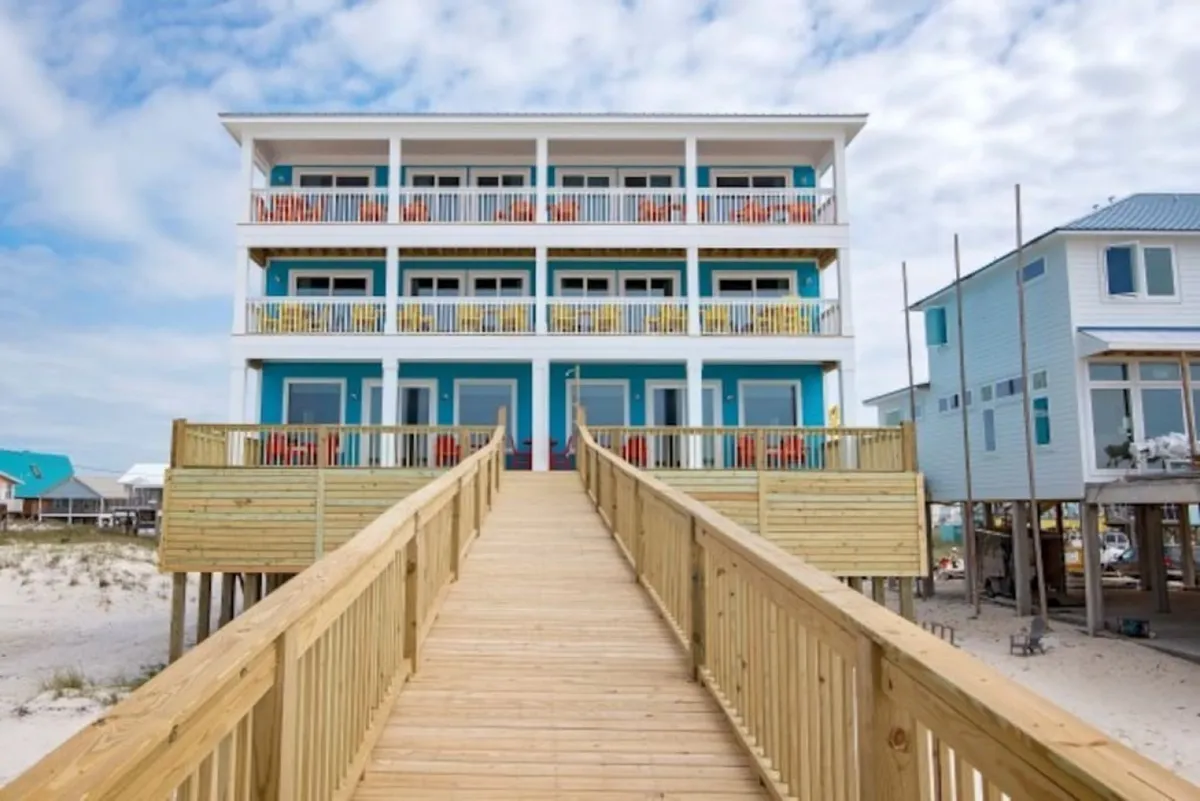 The hero image showcases a striking three-story beach house painted in bright blue and white, with expansive balconies on every level perfect for gathering and enjoying panoramic Gulf views. A wide wooden walkway leads guests from the sandy beach to the inviting entrance, with glimpses of the sugar-white shoreline and nearby homes framing a classic coastal scene.