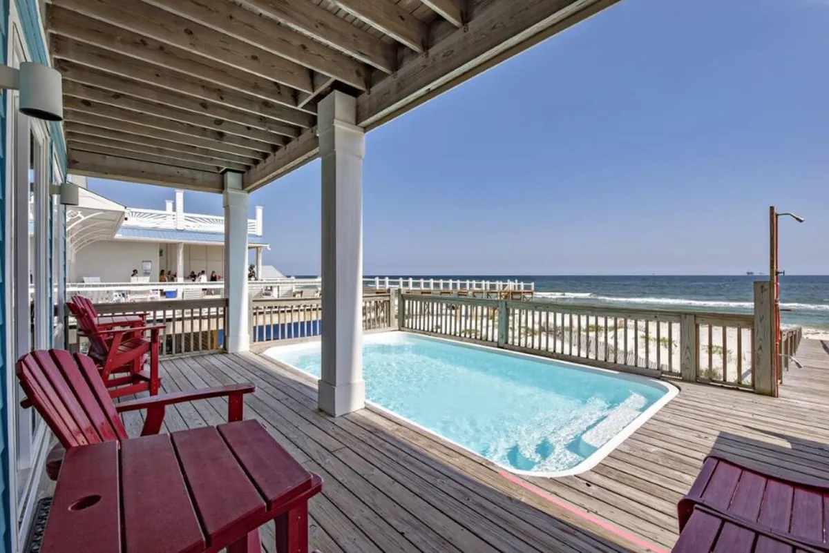 The exterior image focuses on the private pool, wooden deck with red chairs, and a stunning backdrop of the Gulf and sandy beach. The deck invites guests to lounge and socialize, while the pool offers a refreshing retreat steps from the main living areas. The scene is tranquil yet inviting, perfect for group relaxation and celebration.