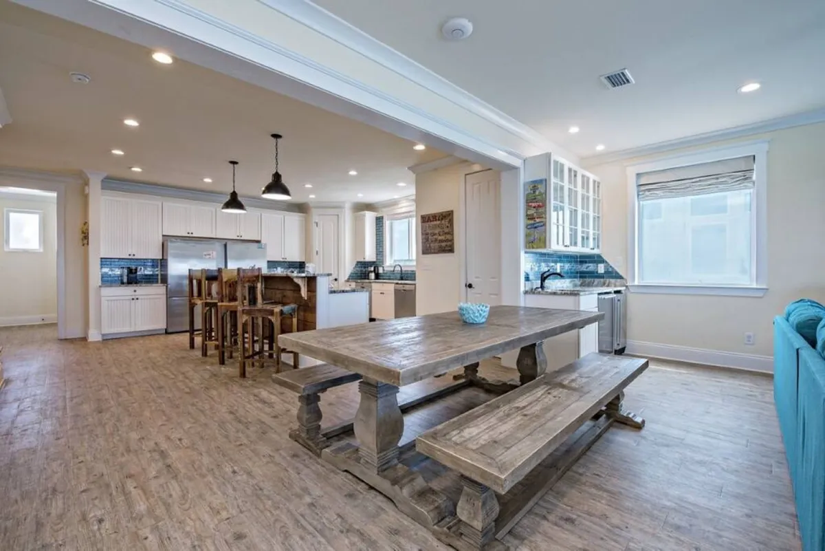 This interior shot highlights a large rustic wooden dining table with benches, a kitchen island with bar stools, and a stainless steel refrigerator. Blue backsplash tiles add a touch of coastal color, and the entire space is bathed in natural light. The layout is open and flows seamlessly between the kitchen and dining area, making it easy for groups to cook, dine, and socialize together.