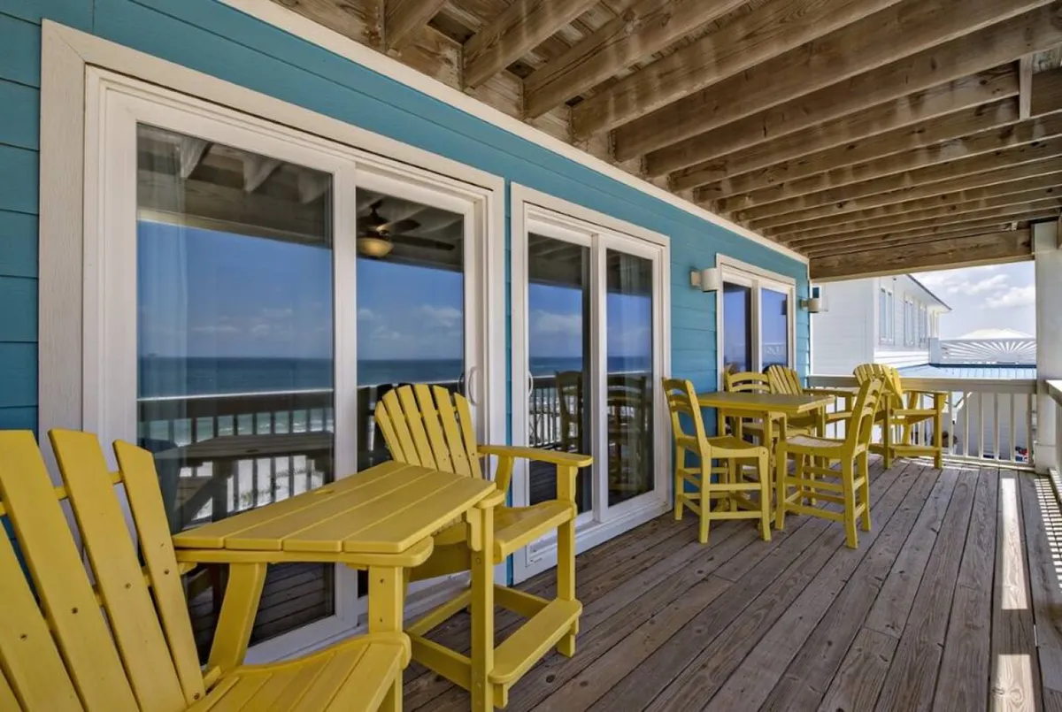 A cheerful balcony scene featuring yellow Adirondack chairs and tables facing the Gulf, with blue house siding, glass sliding doors, and a wooden deck. The space is inviting for outdoor dining, socializing, and soaking in panoramic ocean views—perfect for group conversations or sunset celebrations.