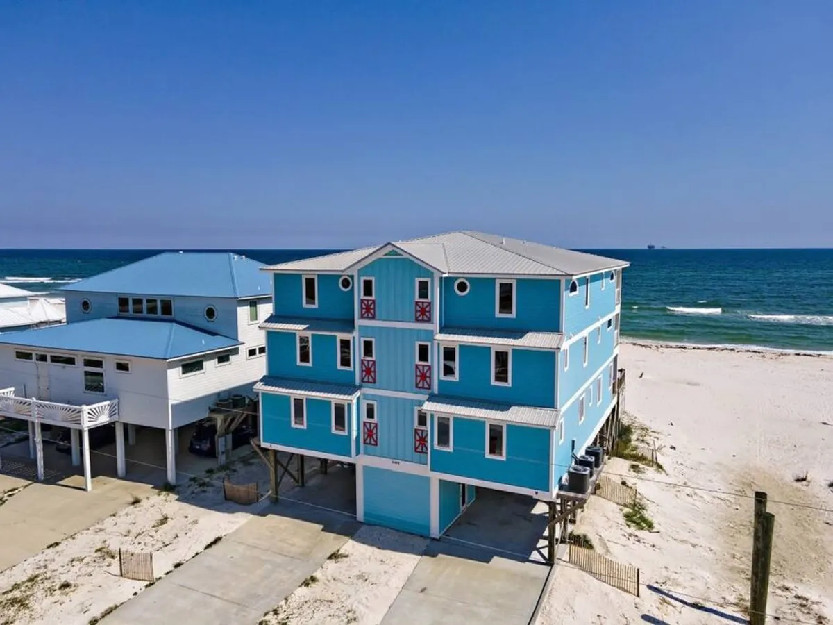 A spacious, turquoise beachfront house with white trim and red accents sits directly on a sugar-white sandy beach, with crystal blue Gulf waters and an adjacent house nearby. The home’s expansive balconies and size evoke the ability to host large groups and social gatherings, while the secluded setting highlights the exclusive, private nature of the property.