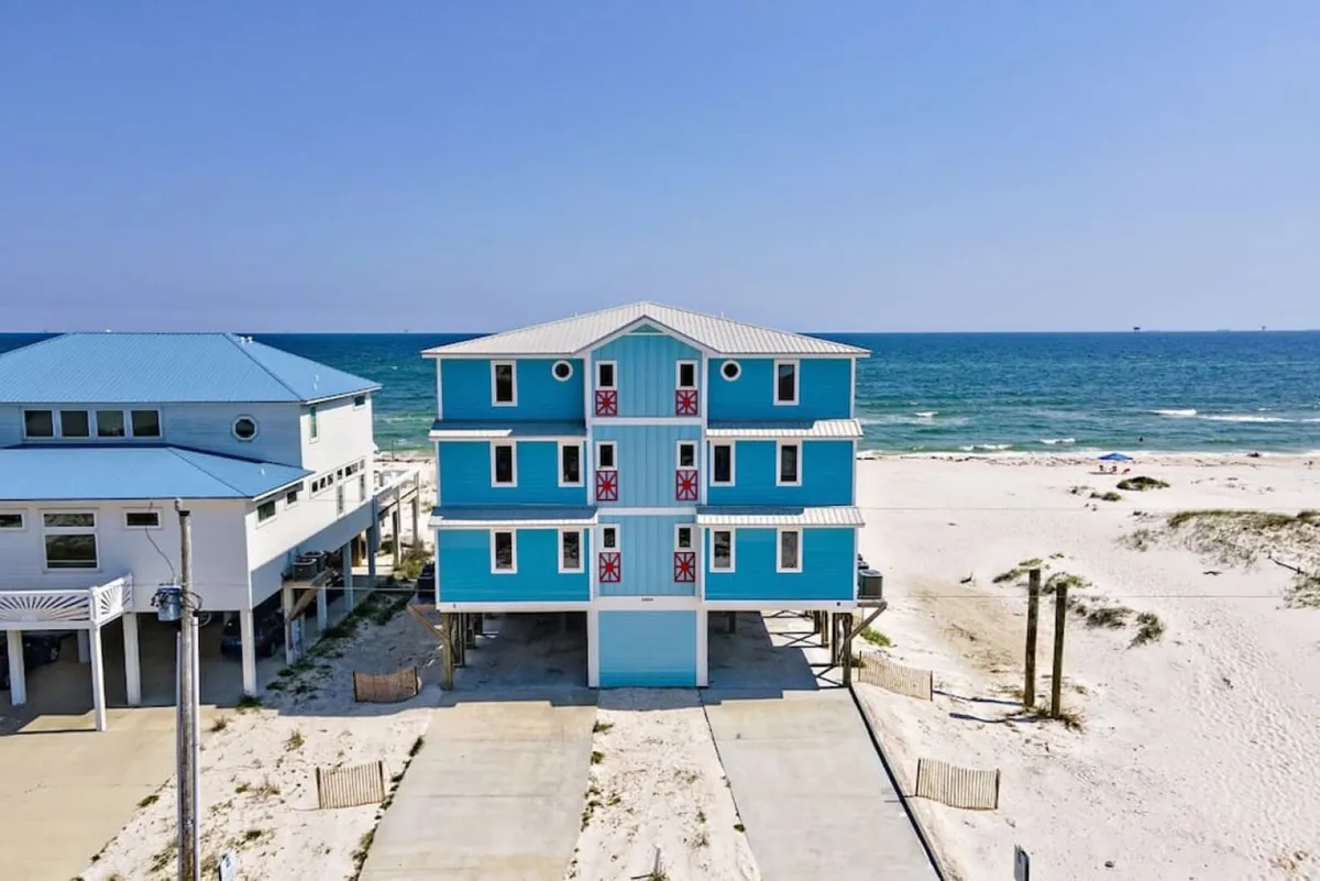 This exterior image showcases a bright blue beach house with red accents set against a pristine white sandy beach and the deep blue Gulf. The house sits near an adjacent coastal home, with the ocean stretching out beyond. The mood is peaceful and rejuvenating, with a luxurious coastal living vibe.
