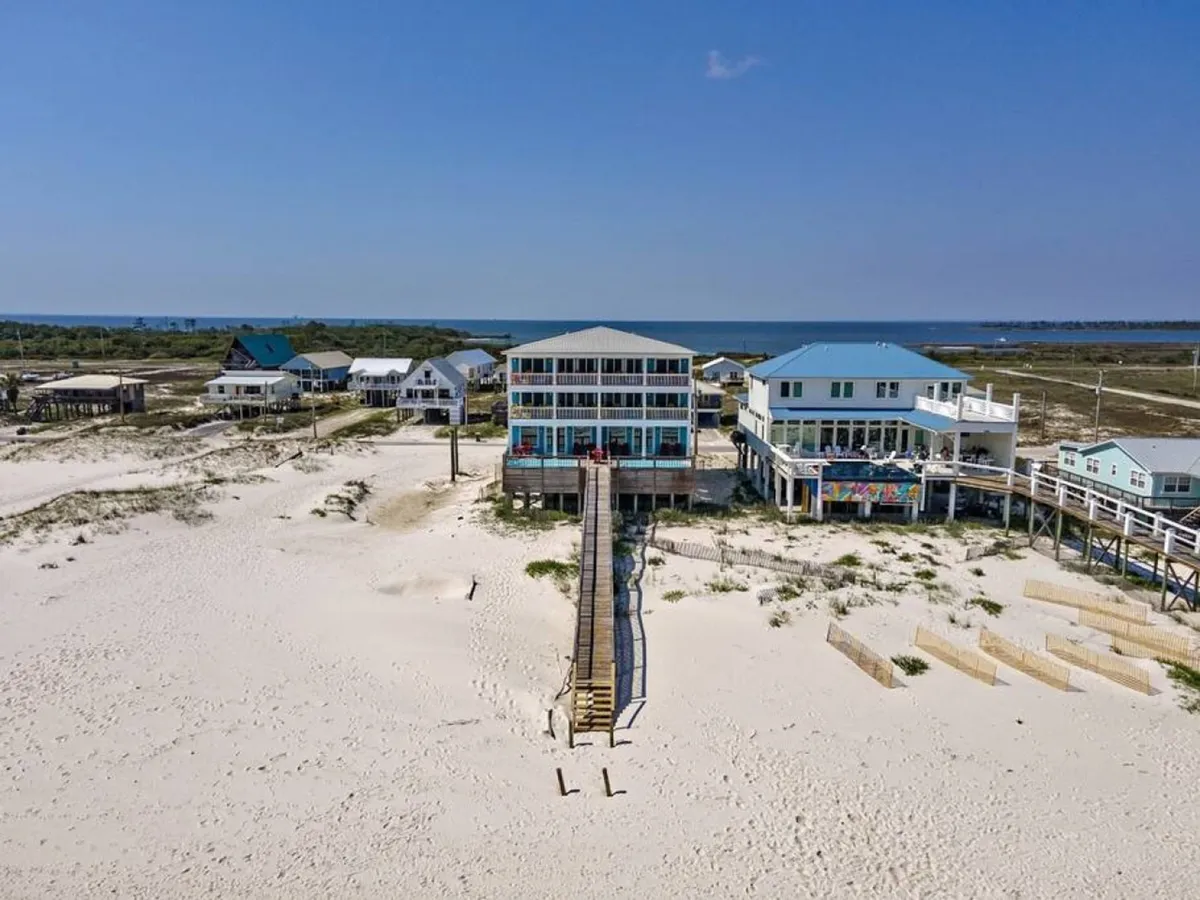 A large beachfront duplex featuring multiple balconies is shown with a long wooden walkway leading directly to a secluded stretch of white sandy beach and natural dunes. Neighboring houses are visible in the background, but the focus is on the home's access to the tranquil shoreline.