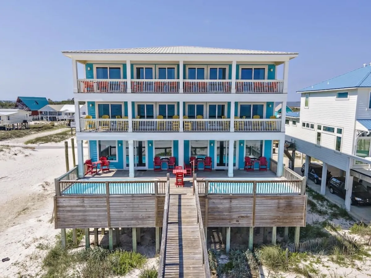 A three-story beachfront house with multiple balconies and a private swimming pool on the ground-level deck. Red chairs are arranged for outdoor relaxation or group activities, and a wooden walkway leads to the beach, suggesting easy access for guests.