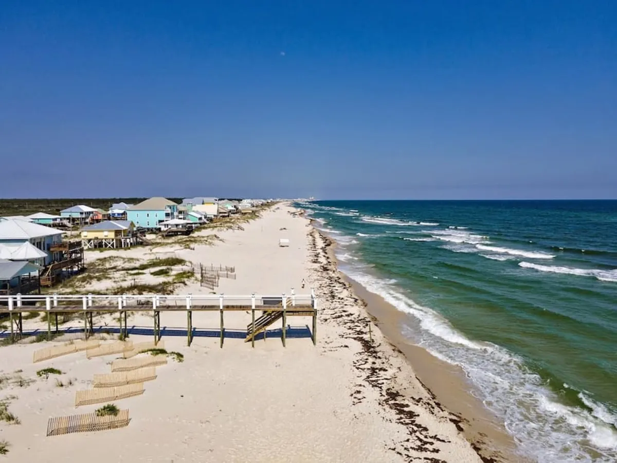 A sweeping view of a pristine white sand beach with a pier extending into the turquoise ocean, and a row of colorful, elevated beach houses lining the shore. The scene is tranquil and inviting, with gently rolling waves and ample open space.