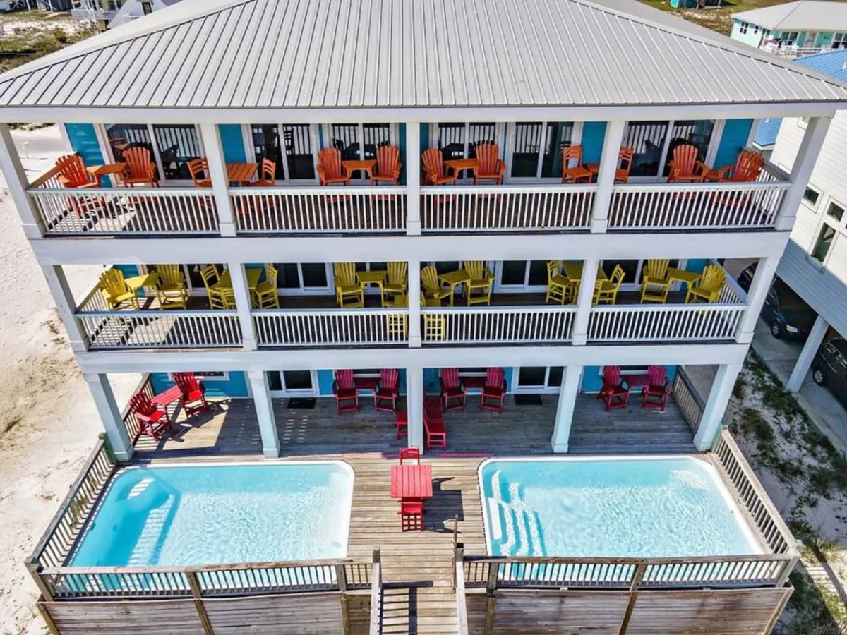 The exterior scene displays a vibrant coastal home with three levels of balconies furnished with colorful chairs and two private swimming pools. The deck area offers plenty of space for outdoor dining, lounging, and socializing, with stunning panoramic views of the Gulf of Mexico in the background.