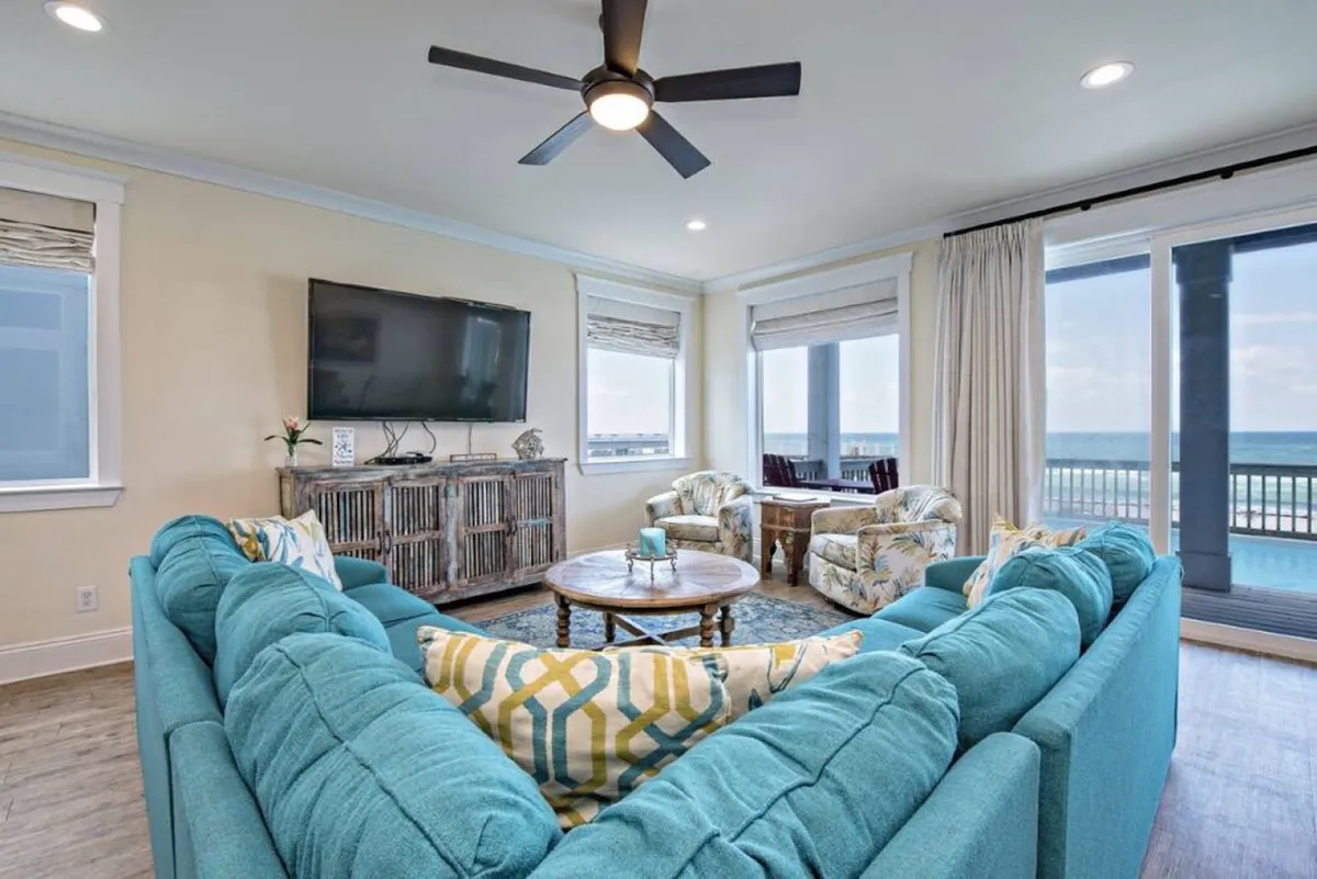 The image showcases a spacious, open-concept living room with a large turquoise sectional sofa, round wooden coffee table, and two floral-patterned armchairs. The room is bright and airy, with large windows and a sliding door revealing views of the ocean and the private pool just outside. The flat-screen TV is mounted above a rustic wooden cabinet, and the entire space exudes a breezy, coastal charm perfect for group gatherings.