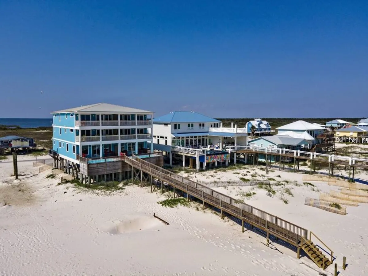 A three-story blue beach house on stilts with wide balconies faces a long wooden boardwalk that leads to the white sandy beach and blue ocean horizon. The scene evokes relaxation and the promise of beachside adventure.