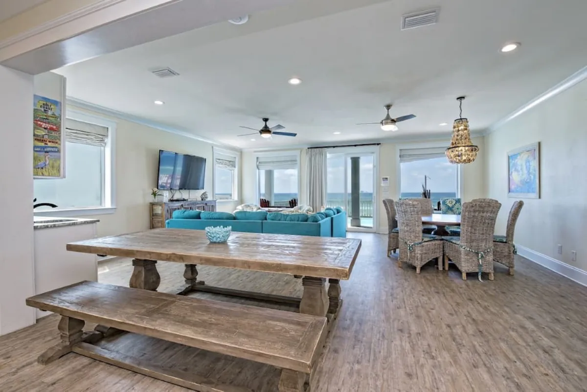 A wide view of the main floor shows a rustic wooden dining table with benches, a turquoise sectional sofa, a flat-screen TV, and woven dining chairs under a chandelier. Through the glass doors, you can glimpse sweeping ocean views. This space is both stylish and comfortable, ideal for family meals, group conversations, and casual lounging after a day at the beach.