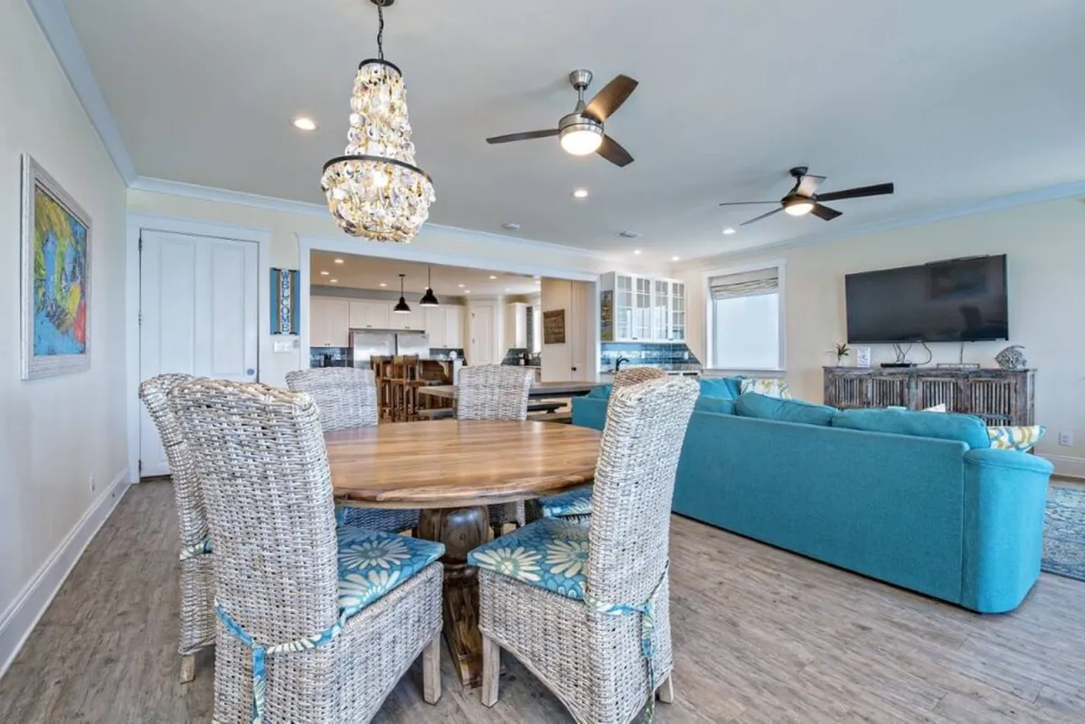 A welcoming open-concept living and dining area with a large turquoise sectional sofa, a round wooden dining table surrounded by wicker chairs, and a chandelier with hanging crystals. The space flows into an open kitchen with bar stools, white cabinetry, and a blue backsplash, all bathed in natural light.