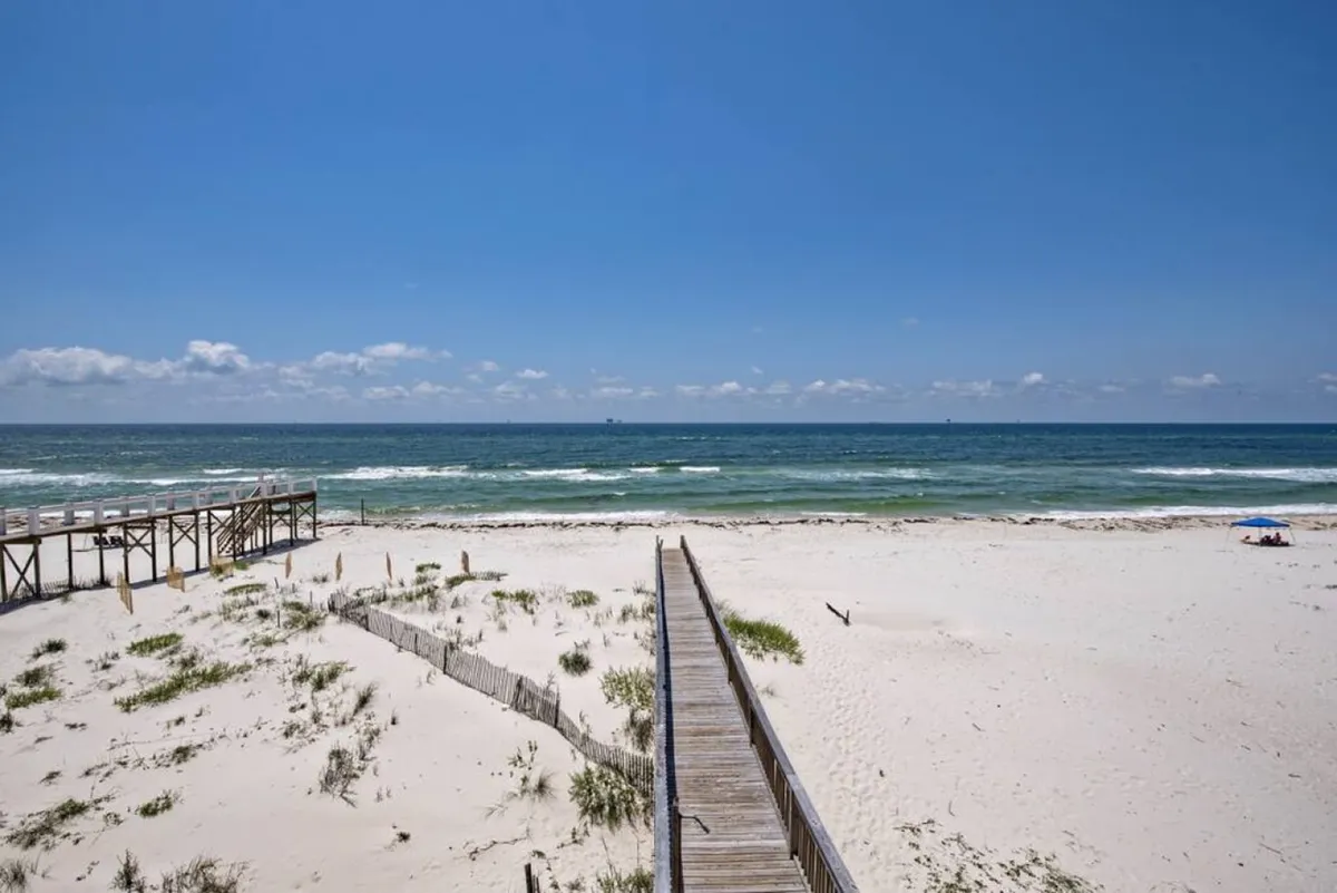 This exterior shot highlights a wooden walkway leading directly to the beach, where white sand meets gentle blue ocean waves. A small shaded umbrella area sits on the sand, underscoring private, tranquil beach access away from crowds.