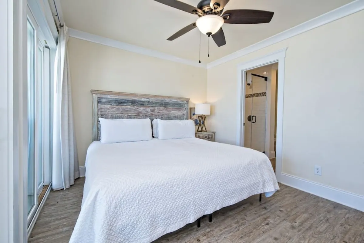 A king-sized bed with a rustic wooden headboard, bedside table with lamp, glass sliding door with sheer curtains leading to a private balcony, and direct access to an en-suite bathroom with a glass shower door. Natural light fills the space, creating a tranquil atmosphere.