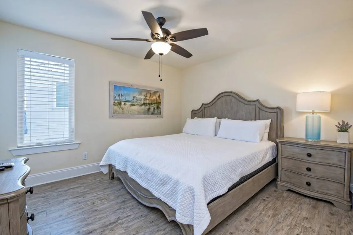A peaceful king-size suite with a gray wooden headboard, blue lamp on a nightstand, a small plant, window with white blinds, and calming beach-themed artwork. The ambiance is soothing, ideal for restful sleep or quiet relaxation.