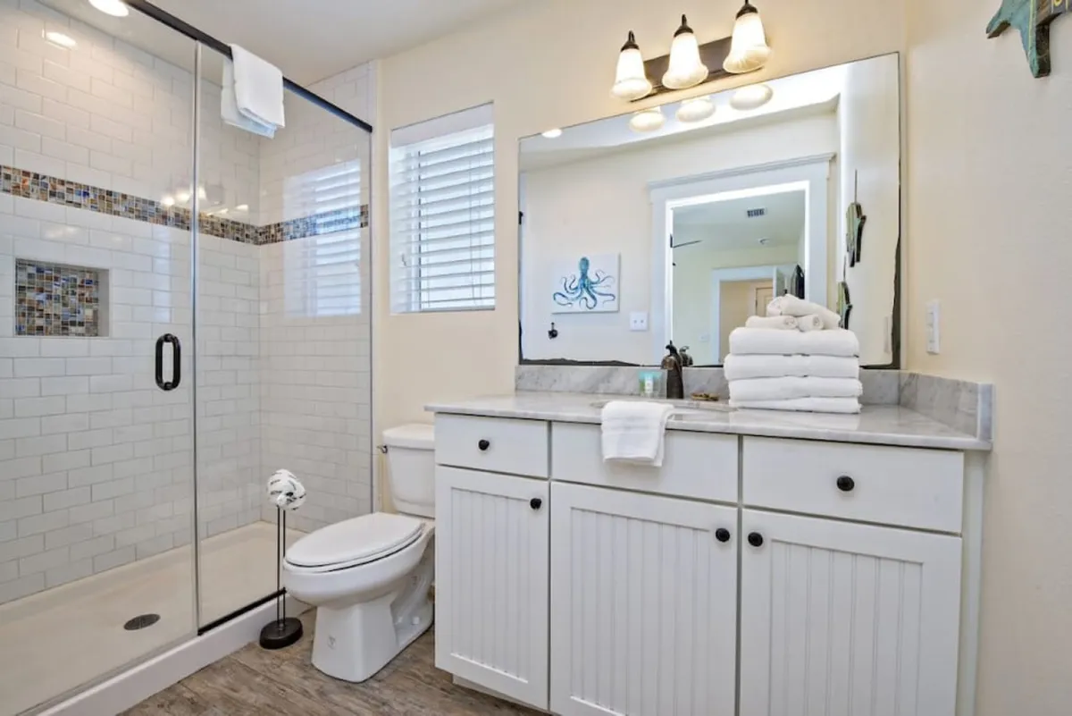 A bright bathroom with a glass-enclosed shower featuring mosaic tile accents, a white vanity with marble countertop, a stack of white towels, and whimsical octopus wall art. The space feels clean, orderly, and refreshing.