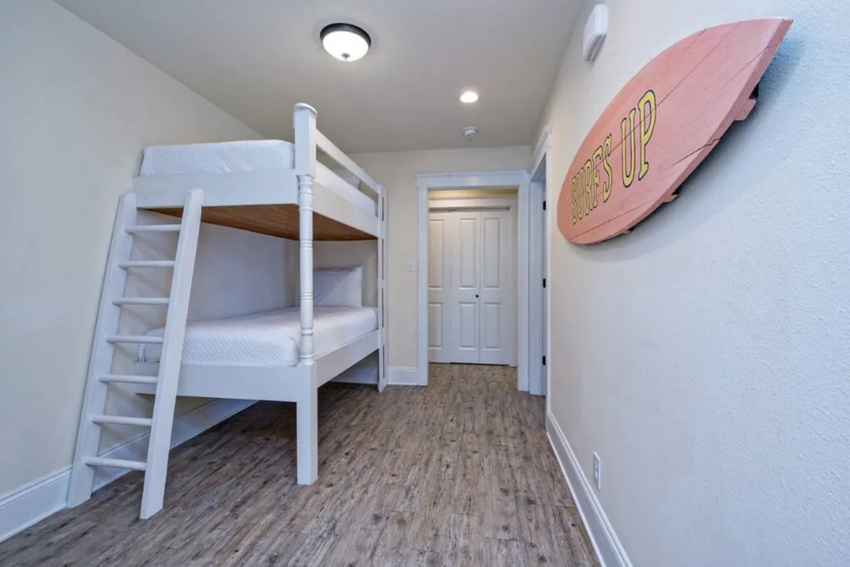 A cozy bunkroom featuring a white wooden bunk bed, playful pink surfboard wall decoration with 'SURF'S UP' text, wood-look laminate flooring, and white trim and closet doors. The space exudes a relaxed, kid-friendly beach vibe.