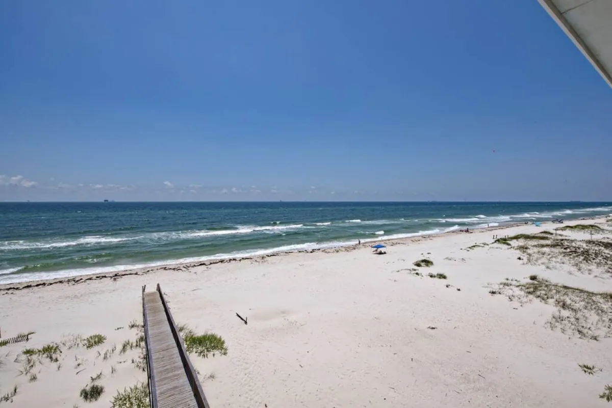 A sweeping view of the sugar-white sandy beach and crystal blue waters of the Gulf of Mexico, with a wooden walkway leading down to the sand. The scene is tranquil and inviting, emphasizing the secluded, private nature of the beachfront setting away from crowds.
