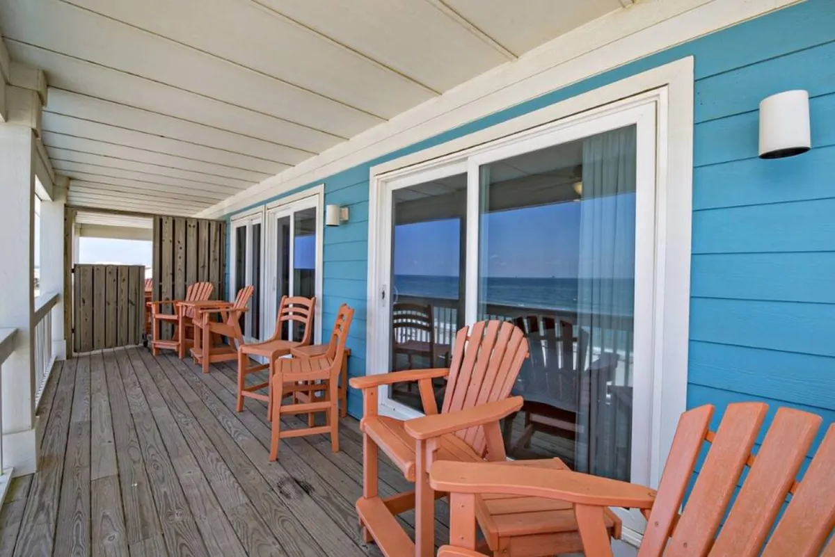 An inviting outdoor balcony with orange Adirondack chairs facing an expansive ocean view. The blue exterior siding and large sliding glass doors add to the coastal charm, while the wooden deck provides ample space for socializing.