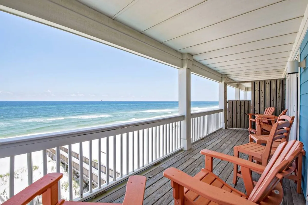This image captures a private balcony with orange Adirondack chairs, a wooden deck, white railing, and a panoramic ocean view framed by the blue exterior wall of the house. The scene is peaceful and open, inviting guests to relax and soak in the fresh Gulf breeze.