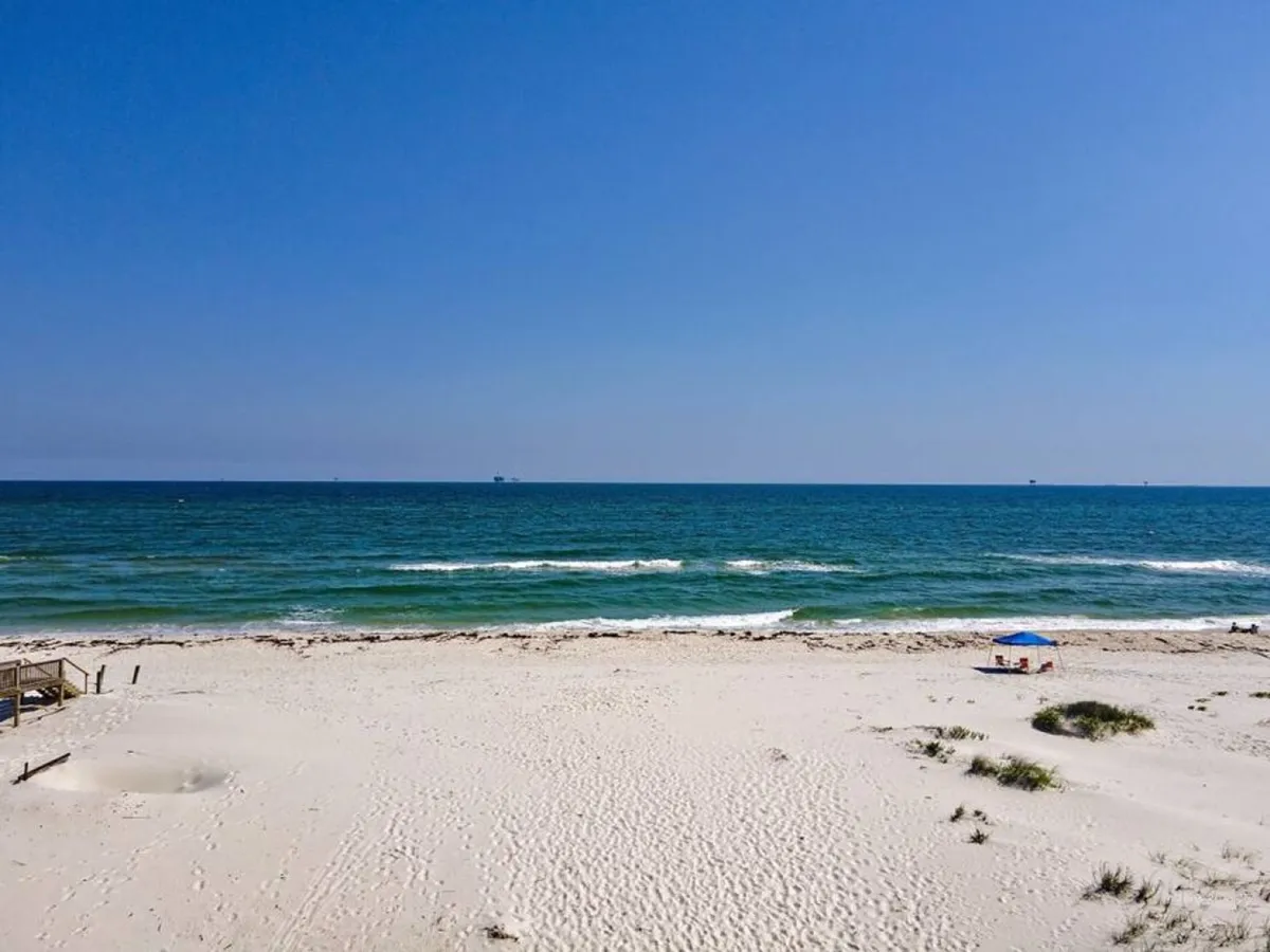 A wide, tranquil beach with sugar-white sand stretches beside crystal blue Gulf waters. A small blue canopy is set up for shade, while distant ships dot the horizon. The scene is open, spacious, and perfect for large groups to spread out and enjoy the sun, surf, and private beach access.